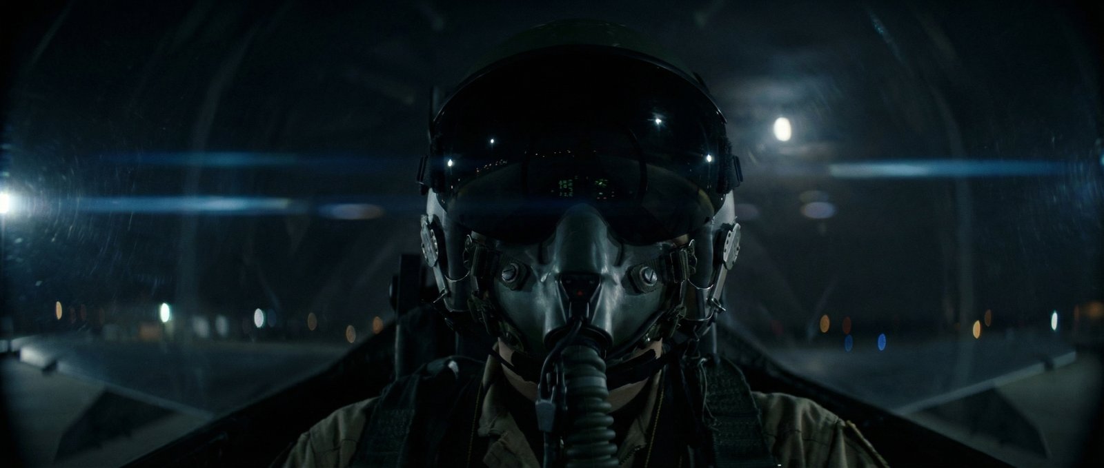 A pilot in a dark cockpit, lit by instruments and a cool blue lens flare.